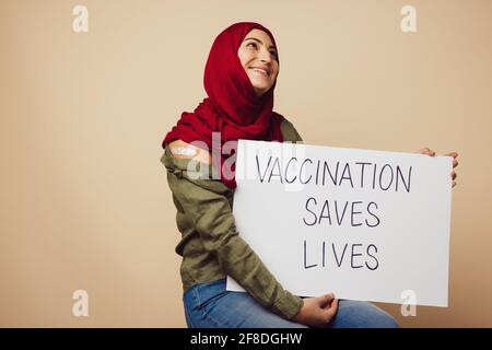 Female wearing hijab holding a banner of 'vaccination saves lives'. Muslim woman after getting vaccine educating people to getting vaccination to save Stock Photo