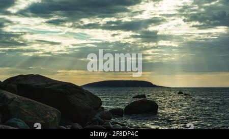 The sun's rays break through the clouds above the sea surface. Seascape with dramatic sky. Shoreline with large boulders in the evening light. Russia, Stock Photo