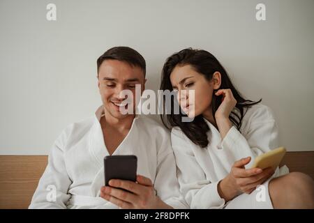Morning of happy young couple in white bathrobes in bed at home, using phones Stock Photo
