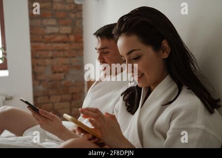 Morning of happy young couple in white bathrobes in bed at home, using phones Stock Photo