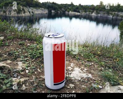 Aluminum beer alcohol or soda or energy drink can mockup 500 ml, isolated on grassy ground against lake surrounded with rocky hill- front view. Drinki Stock Photo