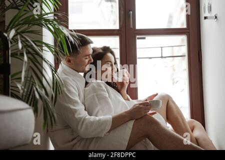 Morning of happy young couple in white bathrobes at home sitting on window, drinking coffee and hug Stock Photo