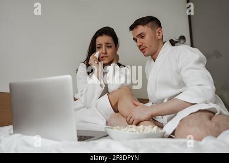 Attractive young couple in bathrobes at home with watching drama movie on laptop, woman is scared Stock Photo
