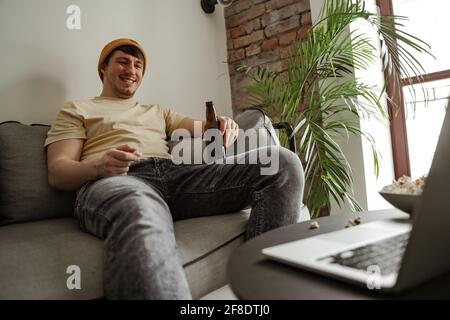 Young man tourist in yellow hat drinking beer and watching on laptop in hotel room, male on vacation. Stock Photo