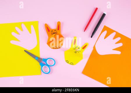 DIY and kids creativity. Colorful paper bunnies from childrens palm of their hand. Greeting cards with bunny on pink background. Gift idea, decor Spring, Easter. Process kid children craft. Stock Photo