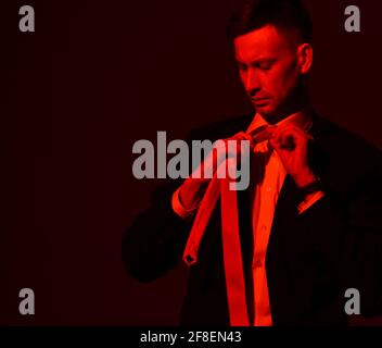 Portrait of young focused businessman in official wear, suit tying his tie looking at it. Red light filter, gel Stock Photo