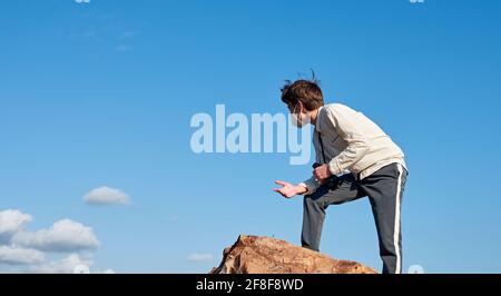 Side view of a Spanish young male wearing a facemask standing on a rock posing against the blue sky Stock Photo