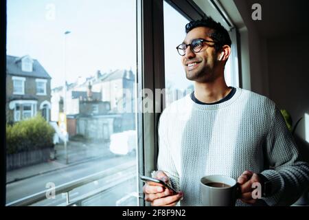 Mixed race business man smiling at window enjoying hot coffee holding cellular device Stock Photo