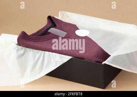 close up blank tag for size, logo, instructions on burgundy sweater in black box and white tissue paper on beige craft paper Stock Photo