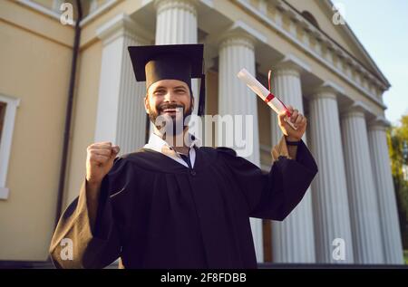 Confident and joyful young man graduate with a diploma in hand shows a gesture of cheers. Stock Photo