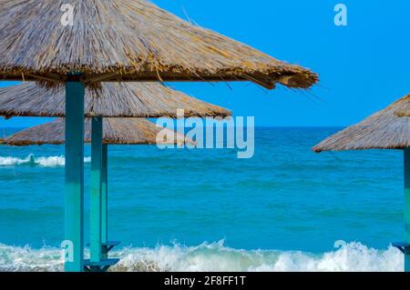 Close-up of straw beach umbrellas providing shade near the wavy water of the Black Sea on sandy shoreline on a sunny clear summer day at Vama Veche, R Stock Photo