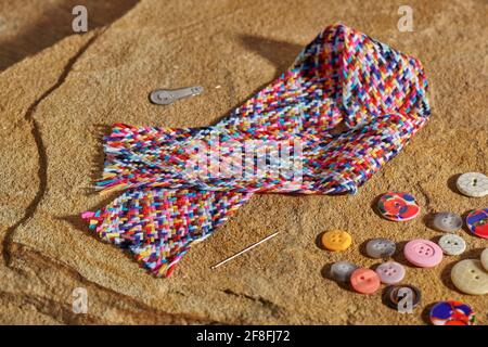 A braid of multi colored sewing threads, needle, needle threader, buttons lying on a stone Stock Photo