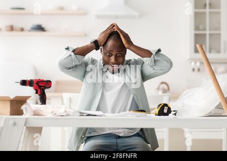 Frightened handyman having problems in assembling new furniture, sitting in kitchen interior Stock Photo