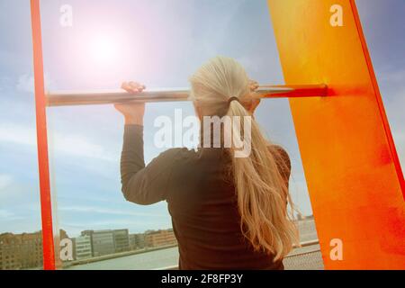Blonde girl doing pulls up on horizontal bar outside by the water.. Woman training doing abs exercises. Stock Photo