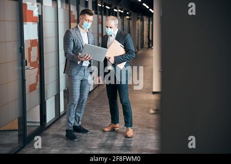 Business colleagues in medical masks talking in office corridor Stock Photo