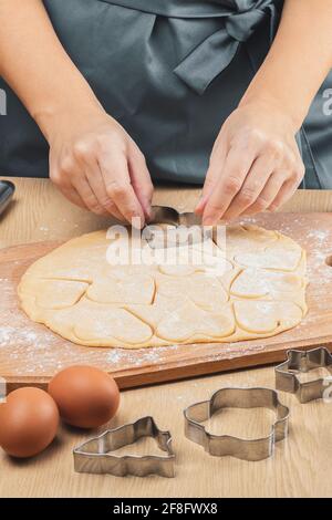 Women's hands hold a metal heart-shaped cookie cutter and cut out cookies from the dough. Close-up, side view. Stock Photo