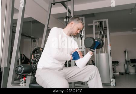 a man in light clothing sitting on a bench lifts dumbbells in the gym. novice bodybuilder Stock Photo