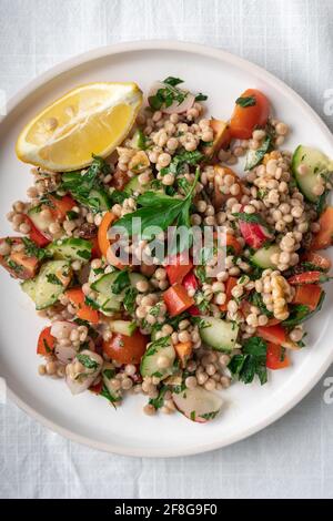 Close-up of a single plate of a healthy vegan wholewheat pearl couscous salad with lemon on white plate and white background, flatlay, vertical