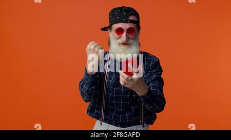 Elderly bearded man use mobile phone browsing online say wow yes, big win news doing winner gesture Stock Photo