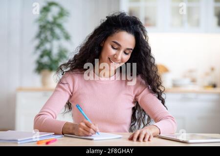 Young Smiling Woman Writing Notes In Notepad, Sitting At Table In Kitchen Stock Photo