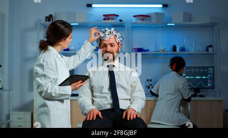 Young man wearing brainwave scanning headset discussing with neurological researcher about symptoms of disease. Medical scientist writing informations of brain scan on tablet working in laboratory Stock Photo
