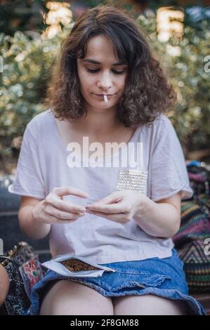 Addicted young woman sitting on park bench and rolling cigarette, preparing to smoke Stock Photo