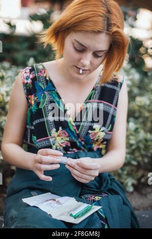 Addicted young woman sitting on park bench and rolling cigarette, preparing to smoke Stock Photo