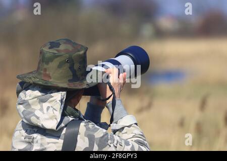professional wildlife photographer, woman in a hat and camouflage suit, camera with a telephoto lens in action Stock Photo