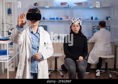 Professional researcher wearing virtual reality glasses using medical inovation in lab analysing brain scan of patient. Team of neurological doctors working with equipment high tech simulator device. Stock Photo