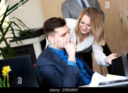 We already have great results! Young beautiful woman pointing at laptop with smile and discussing something with her coworker while standing at office Stock Photo
