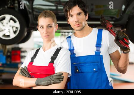 Young woman and man in blue overall - mechanic - stand with in front of a jacked car in a service station Stock Photo