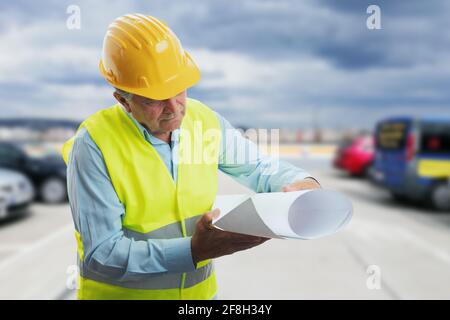Focused old male architect contractor engineer in vest and yellow protective helmet equipment looking at sketches on paper as working concept with par Stock Photo