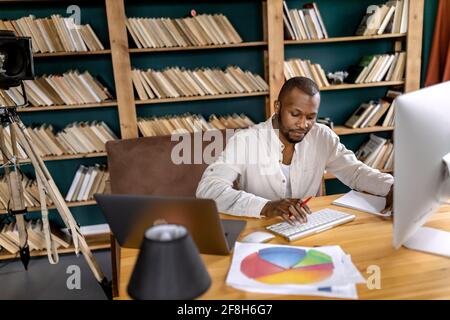 Concentrated african american freelancer sitting at the desk in home office, using computer, chatting with colleagues. Serious businessman doing Stock Photo