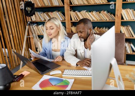 Multiethnic colleagues doing some paperwork, learning financial graphs. Stylish business partners discussing new ideas for project, using computer Stock Photo