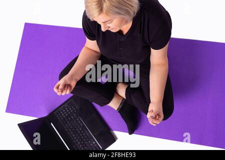 Senior healthy calm woman blonde hair in sportswear meditate watch online class on laptop at home. Fit sporty elderly doing yoga training fitness Stock Photo