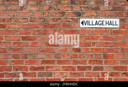 Small black and white Village Hall sign with arrow mounted on a red brick wall. Stock Photo