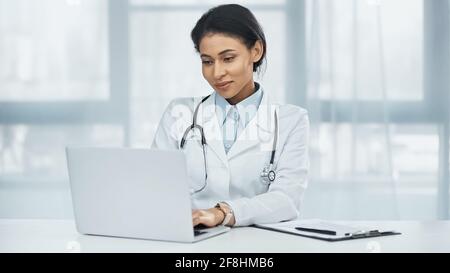 smiling african american doctor using laptop near clipboard on desk Stock Photo