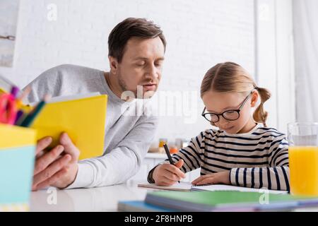 father holding book near daughter writing in notebook while doing homework, blurred foreground Stock Photo