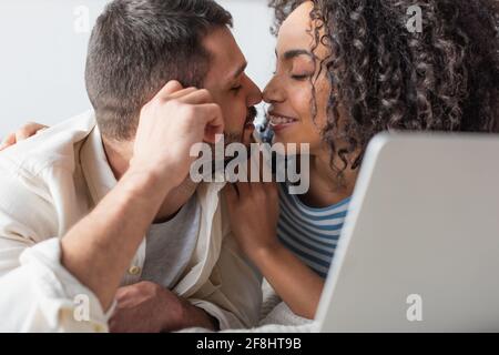 interracial couple kissing while chilling on bed near laptop Stock Photo