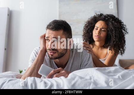 upset man lying on bed and holding smartphone near worried african american woman on blurred background Stock Photo