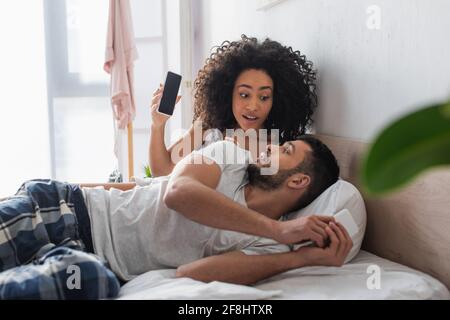 surprised african american woman looking at boyfriend while holding smartphone with blank screen in bedroom