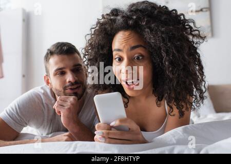 surprised african american woman holding smartphone near bearded boyfriend on blurred background