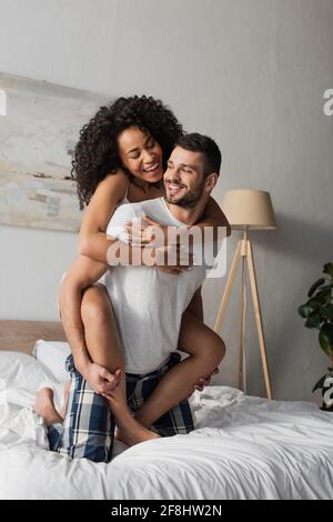 cheerful bearded man piggybacking african american girlfriend in bedroom Stock Photo