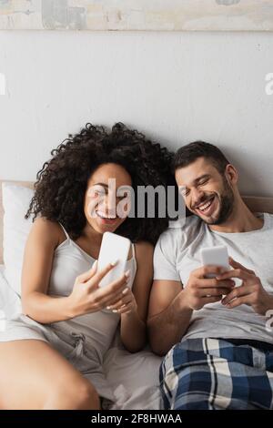 cheerful interracial couple using cellphones and laughing in bedroom Stock Photo