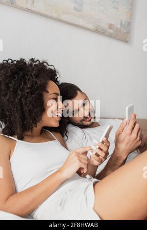 cheerful interracial couple using cellphones in bedroom Stock Photo