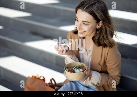 Pretty woman have outdoor lunch near office building while sitting on the stairs and smile. Healthy meal and coffee for takeaway.  Stock Photo