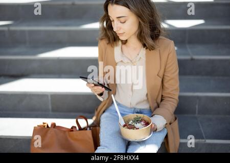 Pretty woman have outdoor lunch near office building while sitting on the stairs and using phone. Healthy meal and coffee for takeaway for workers.  Stock Photo