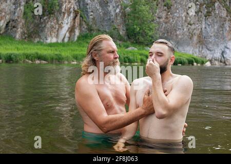 Two bearded men stand in waist-deep water young man with eyes closed and nose pinched by hand listens of older man who holds hands on his chest and ba Stock Photo
