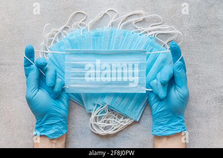 Mens hands in medical gloves holding medical protective mask against the background of masks stack Stock Photo