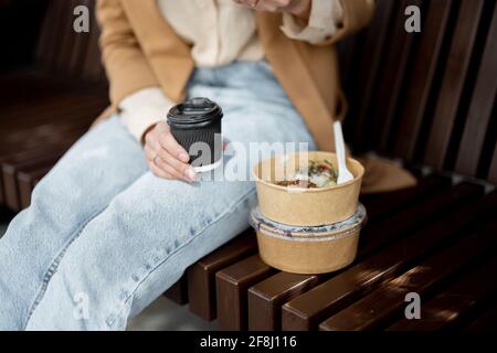 Pretty woman have outdoor lunch near office building while sitting on the bench. Healthy meal and coffee for takeaway for workers. Close up Stock Photo